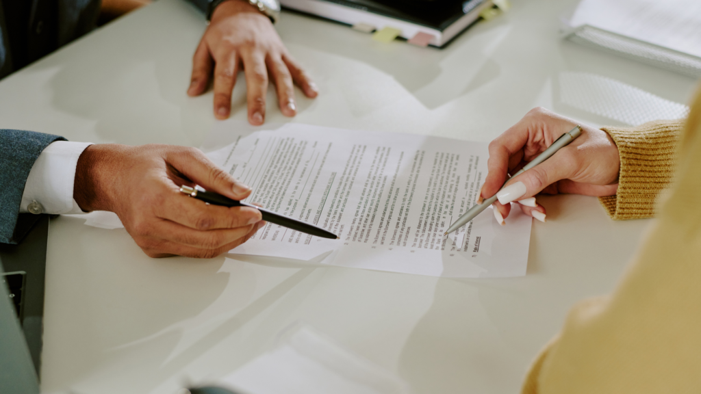 A closeup of a man's hands holding a pen and pointing to a legal document. A woman's hand is also shown holding a pen on the legal document.